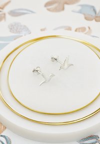 Silver fågelformade örhängen med detaljerade vingar, monterade på en liten pinne, som vilar på en rund vit tallrik med en guldrand.