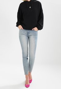 Sudadera negra con capucha y bolsillo frontal, jeans skinny de lavado claro y zapatos de tacón alto de color rosa brillante, exhibidos en una persona de pie.