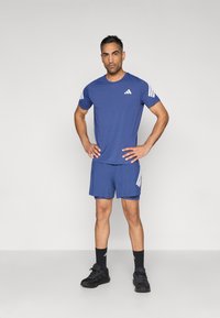 Conjunto deportivo de camiseta y pantalones cortos azules, confeccionado con material ligero. Presenta rayas blancas en los hombros y pantalones cortos a juego. Zapatillas deportivas negras.