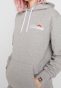 Felpa grigia con cappuccio dotata di tasca a marsupio, polsini elasticizzati e un logo ricamato bianco con un accento colorato. Tessuto morbido e dalla texture strutturata.