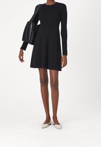 Femme portant une robe noire à manches longues, des ballerines blanches pointues, et tenant un grand sac à bandoulière noir sur un fond blanc uni.