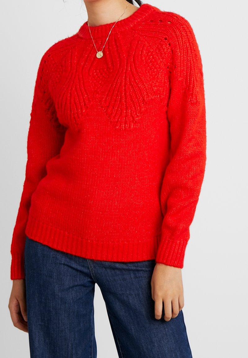 Femme portant un pull en laine rouge vif avec des motifs texturés sur la poitrine et les manches, assorti à un jean bleu foncé et un collier pendentif en or.