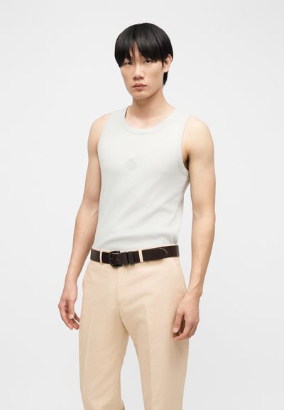 Νεαρός άνδρας με ίσια μαύρα μαλλιά, φορώντας λευκό αμάνικο φανελάκι, μπεζ παντελόνι και μαύρη ζώνη, στέκεται μπροστά σε απλό φόντο.