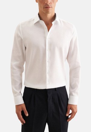 Camisa blanca de manga larga con botón y cuello clásico, que presenta una sutil textura y un detalle en el interior del cuello en contraste. Combinada con pantalones oscuros.