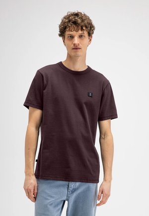 Jonge man met krullend haar, die een effen donkerpaarse korte mouw T-shirt en lichtblauwe spijkerbroek draagt, staand tegen een witte achtergrond.