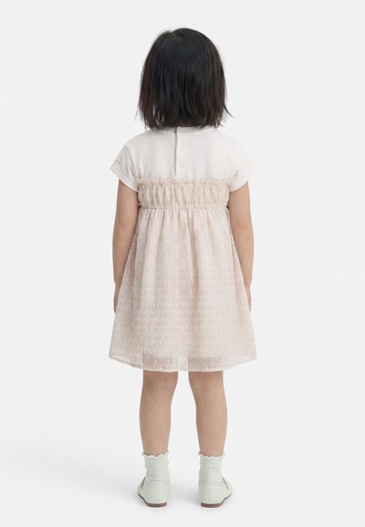 Παιδί με κοντά σκούρα μαλλιά που φοράει ένα ανοιχτό ροζ φόρεμα με σφικτή μέση, λευκές κάλτσες και λευκά παπούτσια, στέκεται γυρισμένο προς τα πίσω σε λευκό φόντο.