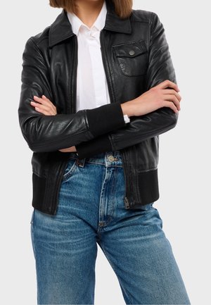 Person iført en sort læderjakke over en hvid skjorte og blå jeans, arme krydset ved livet, med en neutral lys grå baggrund.