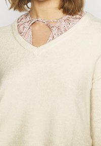 Kremowy sweter z dekoltem w serek i miękką fakturą, nałożony na jasnoróżową bluzkę z wzorem w kropki i wiązaniem.