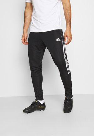 Mann trägt schwarze Adidas-Trainingshose mit weißen Streifen, schwarze Fußballschuhe mit gelbem "NEMESIS"-Schriftzug und ein weißes T-Shirt.