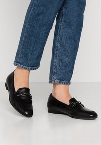 Svarta läderloafer med låg klack, rund tå och dekorativ metalldetalj. Bärs med blå denim i en kortad längd.