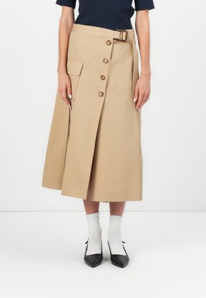 Beige midi-rok met knopen aan de voorkant en een zijzak, gedragen met zwarte puntige schoenen en witte sokken, gecombineerd met een donker geribd topje.