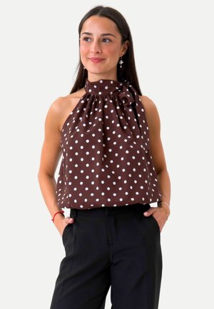 Donna con top marrone a pois senza maniche con fiocco al collo e pantaloni neri, mani in tasca, guarda leggermente di lato sorridendo.