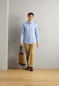 Chemise à manches longues bleu clair, pantalon beige, chaussures marron, tenant un sac fourre-tout marron et noir, debout sur un sol en bois contre un mur gris.