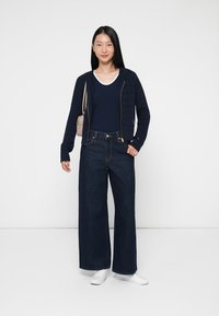 Granatowy sweter zapinany na zamek, granatowy T-shirt, wysoko taliowane ciemnoniebieskie spodnie typu wide-leg oraz białe trampki tworzą swobodny wygląd. Mała torebka.