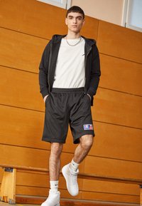 Sudadera negra con cremallera sobre una camiseta blanca, combinada con pantalones cortos de malla negra con un parche de logo. Zapatillas blancas y de pie sobre un banco.