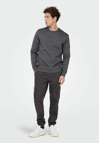 Jeune homme en sweat-shirt gris foncé et pantalon cargo avec des baskets blanches, debout et regardant sur le côté sur un fond clair uni.
