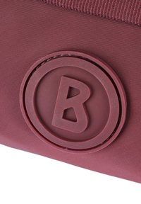 Bordová taška s texturovaným povrchem, kulatým gumovým logem se vyvýšeným "B" uprostřed a zpevněným šitím podél okrajů.