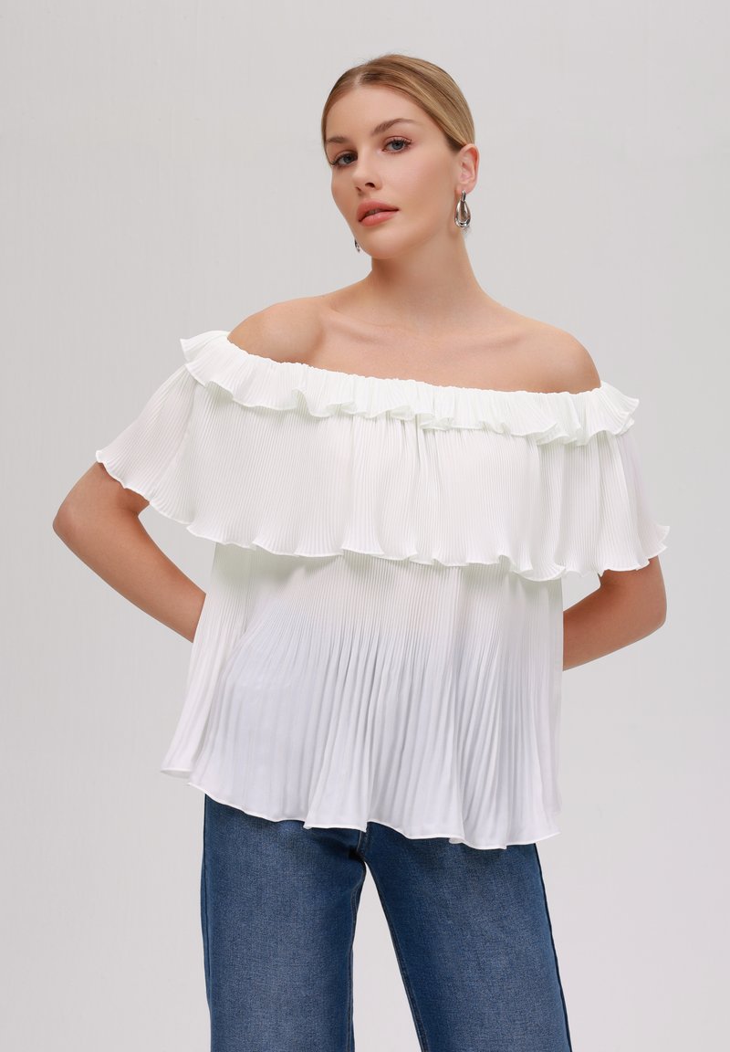 Weiße schulterfreie Bluse mit Rüschensaum, gepaart mit einem plissierten Stoff und einem lässigen Schnitt, kombiniert mit blauen Jeans.