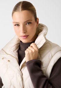 Gesteppte cremefarbene Steppweste mit hohem Kragen, goldenen Akzenten und Reißverschluss. Über einem dunkelbraunen Rollkragenpullover getragen. Glatte Textur.