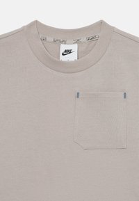 Pelēks kokvilnas T-krekls ar apaļu apkakli, vienu krūšu kabatu un kontrastējošem šuvēm. Iekšējā apkakles tagā ir norādīts "Nike". Mīksts tekstūras materiāls.