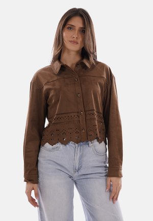 Donna che indossa una giacca di camoscio marrone con bottoni, decorata con motivi intagliati al laser all'orlo, e jeans azzurri a vita alta, in piedi su sfondo bianco.