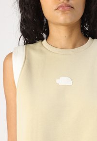 Beige ärmlös topp med ribbad halsringning, som har en vit broderad logotyp på bröstet och kontrasterande vita axelremmar.
