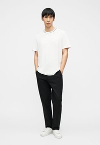 Junger Mann steht mit den Händen in den Taschen, trägt ein weißes T-Shirt, schwarze Hose und weiße Turnschuhe vor einem schlichten hellen Hintergrund.