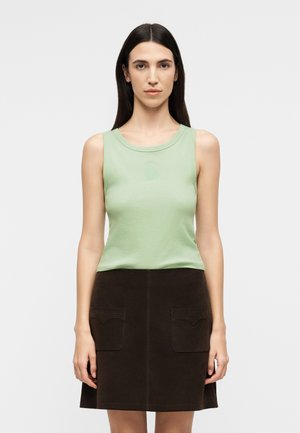 Γυναίκα με μακριά σκούρα μαλλιά, που φοράει αμάνικο ανοιχτό πράσινο τοπ και σκούρη καφέ φούστα με εμπρόσθιες τσέπες, στέκεται μπροστά σε απλό φόντο.