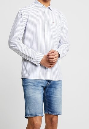 Mężczyzna w białej koszuli z cienkimi ciemnymi paskami i jasnoniebieskich denimowych szortach, stojący ze splecionymi rękami.