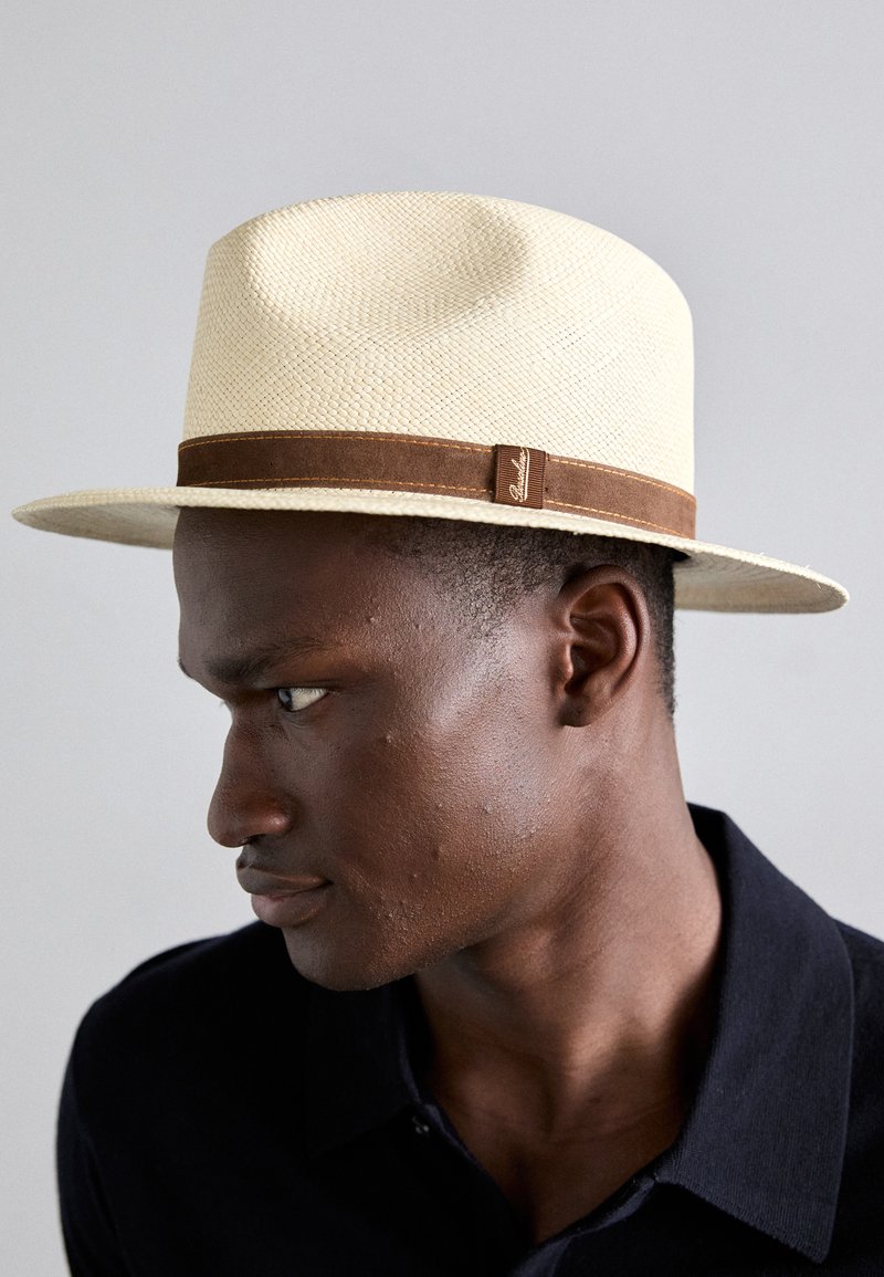 Bēšas salmu fedora cepure ar brūnu auduma joslu un smalku logotipa detaļu, ar plašu malu un teksturētu apdari.