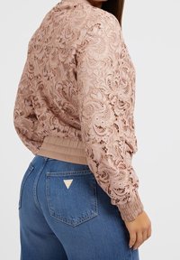 Blouson aviateur en dentelle rose clair avec un motif floral et des poignets côtelés, porté avec un jean en denim bleu foncé. Logo de marque notable sur le jean.