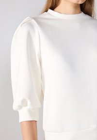 White sweatshirt featuring a high neckline, puffed sleeves, and smooth texture. The cuffs are fitted, and the fabric appears soft and lightweight.