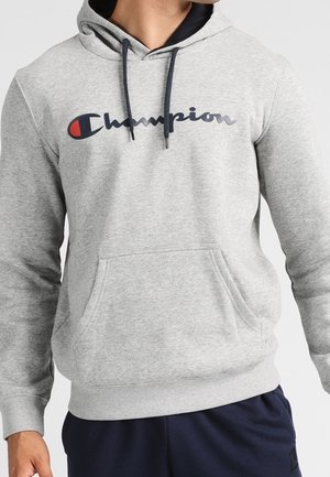 Mann trägt einen hellgrauen Champion-Hoodie mit Fronttasche und marineblauen Kordeln, kombiniert mit dunkelblauen Hosen.