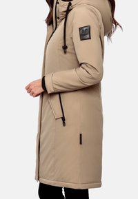 Cappotto lungo beige con cappuccio, dotato di accenti neri, tasche con zip e una patch con logo sulla manica. Materiale liscio, dettagli cuciti.