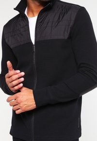 Černá bunda na zip s texturovaným nylonovým horním panelem a žebrovanou pletenou dolní částí. Dlouhé rukávy mají jemné detaily a lichotivý střih.