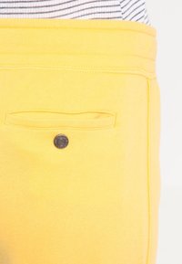 Žluté tepláky s viditelnou zadní kapsou, která má černý knoflík. Proužkovaná látka je částečně vidět nahoře. Hladký povrch.