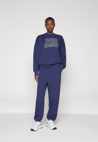 Sötétkék pulóver és hozzáillő nadrág bordás mandzsettákkal. A pulóveren világos színű "UNDER ARMOUR" felirat található. Sportcipők fehérek.