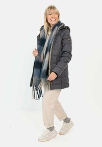Gezimde zwarte gewatteerde parka met capuchon, twee voorzakken en een bruin logo-opdracht. Blauw en crème gestreepte sjaal met franjes. Draagt beige broeken en sneakers.