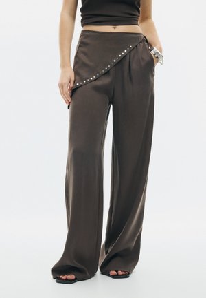 Žena v hnedých širokých nohaviciach s asymetrickým zábalovým detailom a čiernom bezrukávovom krátkom topu, stojaca s jednou rukou v vrecku.