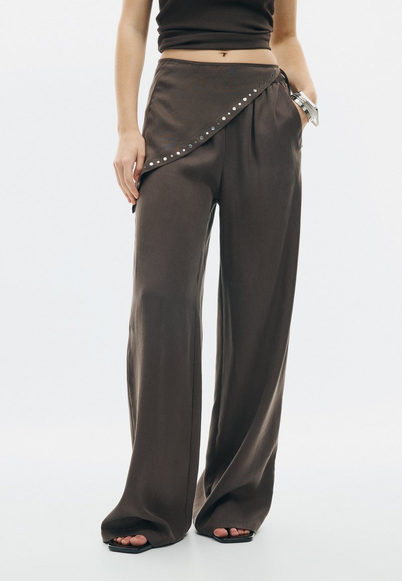 Femme vêtue d'un pantalon marron à jambes larges avec un détail noué asymétrique et un crop top noir sans manches, debout avec une main dans la poche.