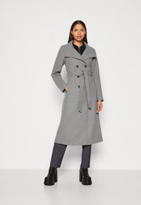 Manteau gris croisé avec une taille ceinturée, des revers larges et des poches latérales. Associé à un pantalon gris foncé et des bottes à talons épais noires.