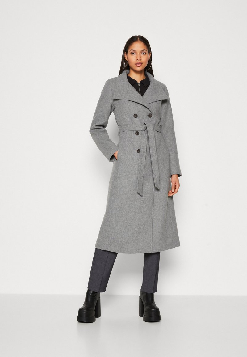 Manteau gris croisé avec une taille ceinturée, des revers larges et des poches latérales. Associé à un pantalon gris foncé et des bottes à talons épais noires.