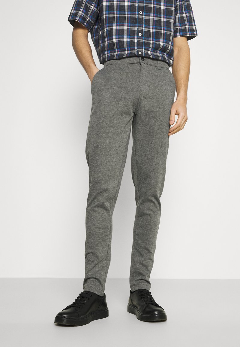 Pantalon gris fuselé en matériau doux, présentant une texture lisse et une finition subtile. Porté avec des baskets noires.
