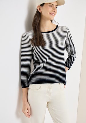 Femme aux longs cheveux bruns portant une casquette beige, un chemisier à manches longues rayé noir et blanc, et un pantalon blanc cassé, debout contre un mur.