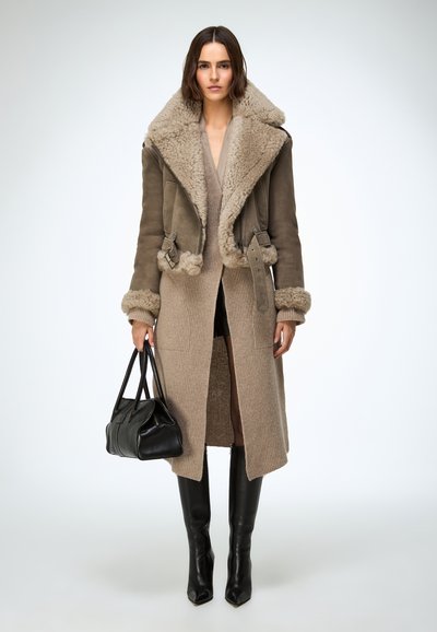Manteau en cuir taupe doublé en peau lainée, avec un col large, des détails à boutons et boucles, porté avec une robe en maille beige et un sac à main noir.