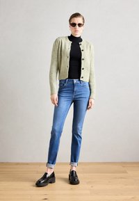Lichtgroene gebreide cardigan met zwarte knopen, gedragen over een zwarte coltrui. Blauwe jeans met opgerolde manchetten en zwarte platform instappers.