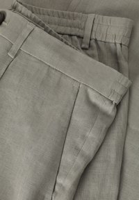 Lichtgrijze linnen broek, opgevouwen, met een platte voorkant, zijzakken en een knoopsluiting. Zachte textuur met fijn gestikte naden.