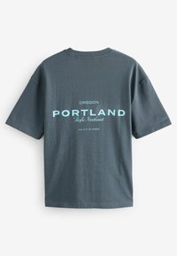 Pelēka kokvilnas t-krekls ar īsām piedurknēm, uz muguras ar gaiši zilu uzrakstu "PORTLAND", kā arī "OREGON" un atrašanās vietas detaļas.