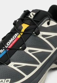 Szürke atlétikai cipő fekete és sárga fűzőrendszerrel, texturált felsőrésszel, fehér részletekkel és feltűnő márkalogóval az oldalon.