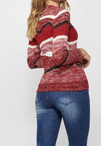 Rot-weißer, gemusterter Strickpullover mit langen Ärmeln und geripptem Saum, kombiniert mit blauen Jeans, die einen leichten Verwaschungseffekt und Gesäßtaschen haben.